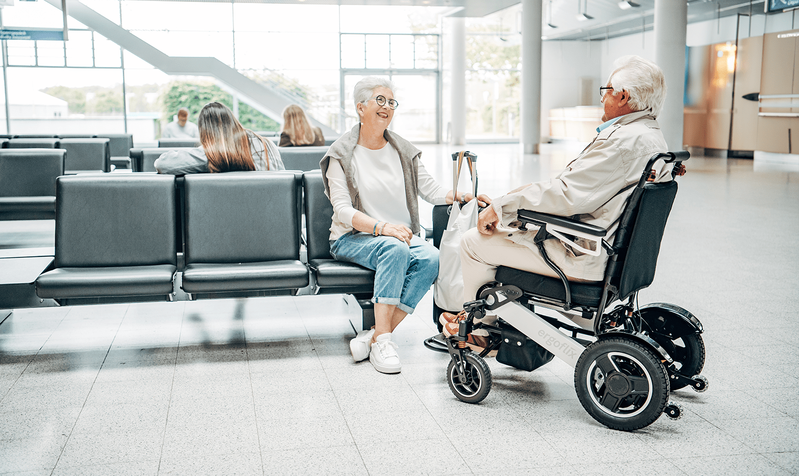 Dieses Bild zeigt einen Mann im faltbaren E-Rollstuhl ergoflix® LX am Flughafen, der mit seiner Frau spricht.
