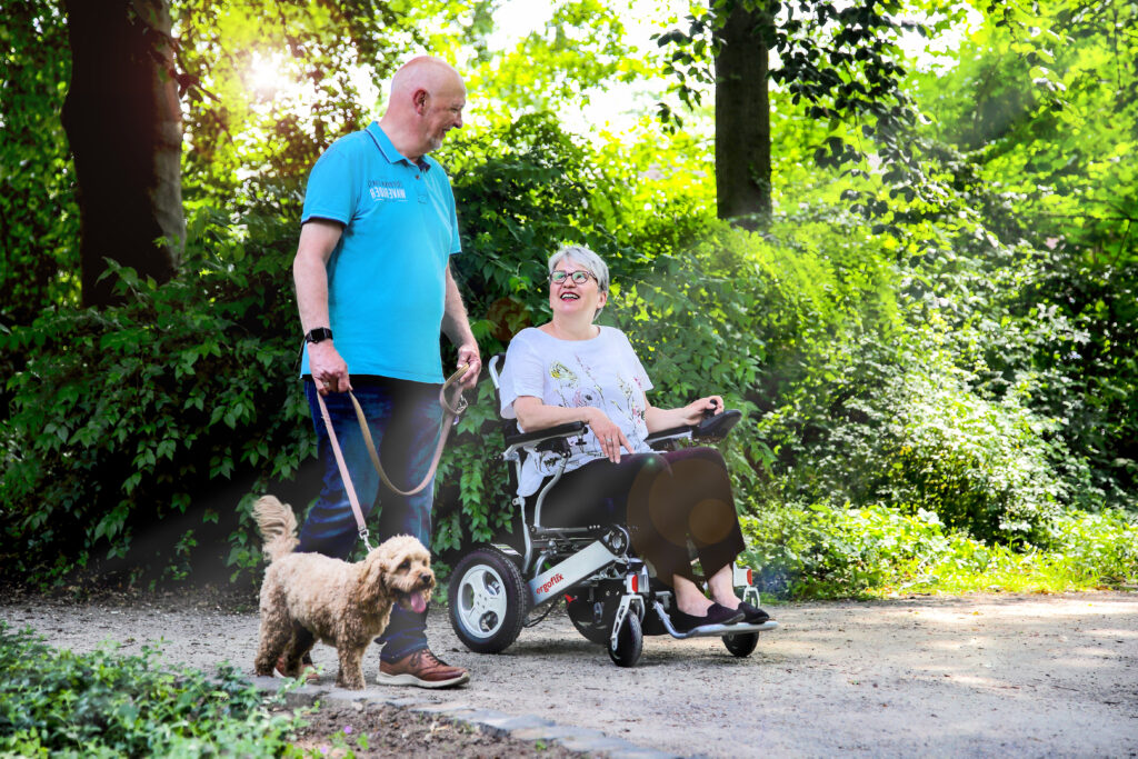 Dieses Bild zeigt einen ergoflix-Rollstuhl, indem eine Frau sitzt. Ein Mann mit Hund läuft daneben.