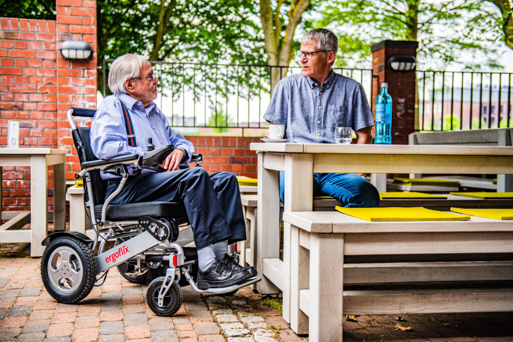 Dieses Bild zeigt einen Herrn im ergoflix-Rollstuhl, wie er mit seinem Freund in der Außengastronomie sitzt